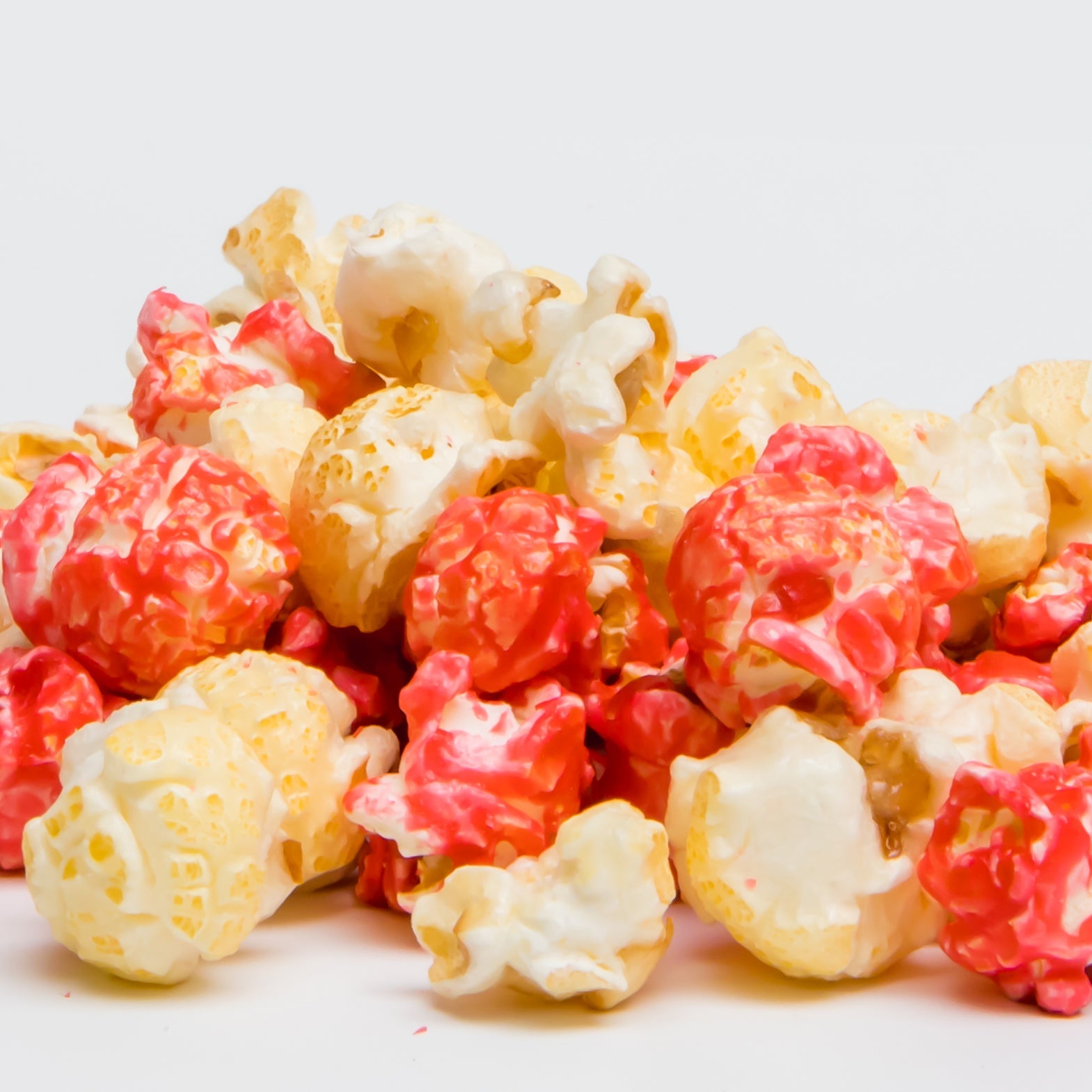 Strawberry Cheesecake Popcorn piles gleam with red and white candy glaze against a white background, making this gourmet treat irresistible to popcorn lovers.
