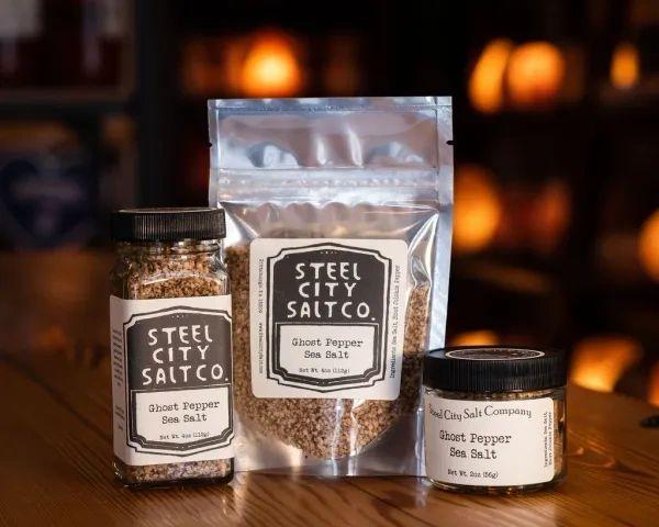 Three containers of Ghost Pepper Sea Salt—a glass jar, resealable pouch, and plastic tub—sit on a wooden surface with matching black-and-white labels. Warm lighting highlights this spicy salt made with Bhut Jolokia (Ghost Pepper).