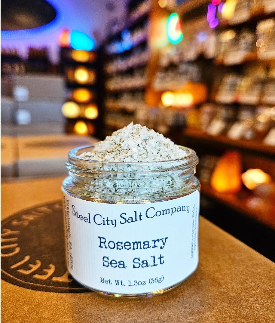 A jar labeled &quot;Rosemary Sea Salt,&quot; a hand-blended salt, sits on a counter in a warmly lit store, with softly blurred shelves and glowing lights in the background.