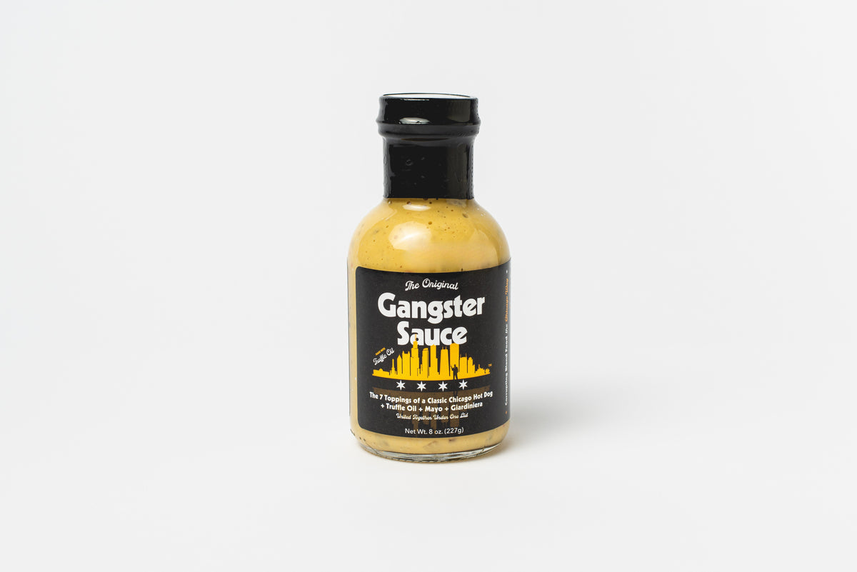 A bottle of Gangster Sauce Chicago hot dog sauce stands upright on a white background, featuring a black label with a yellow city skyline and white text.