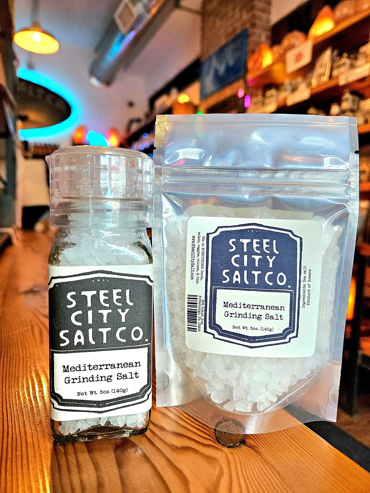 Two containers of Mediterranean Grinding Salt sit on a wooden counter: one is a grinder and the other a resealable bag, with blurred shelves and colorful lights in the background.