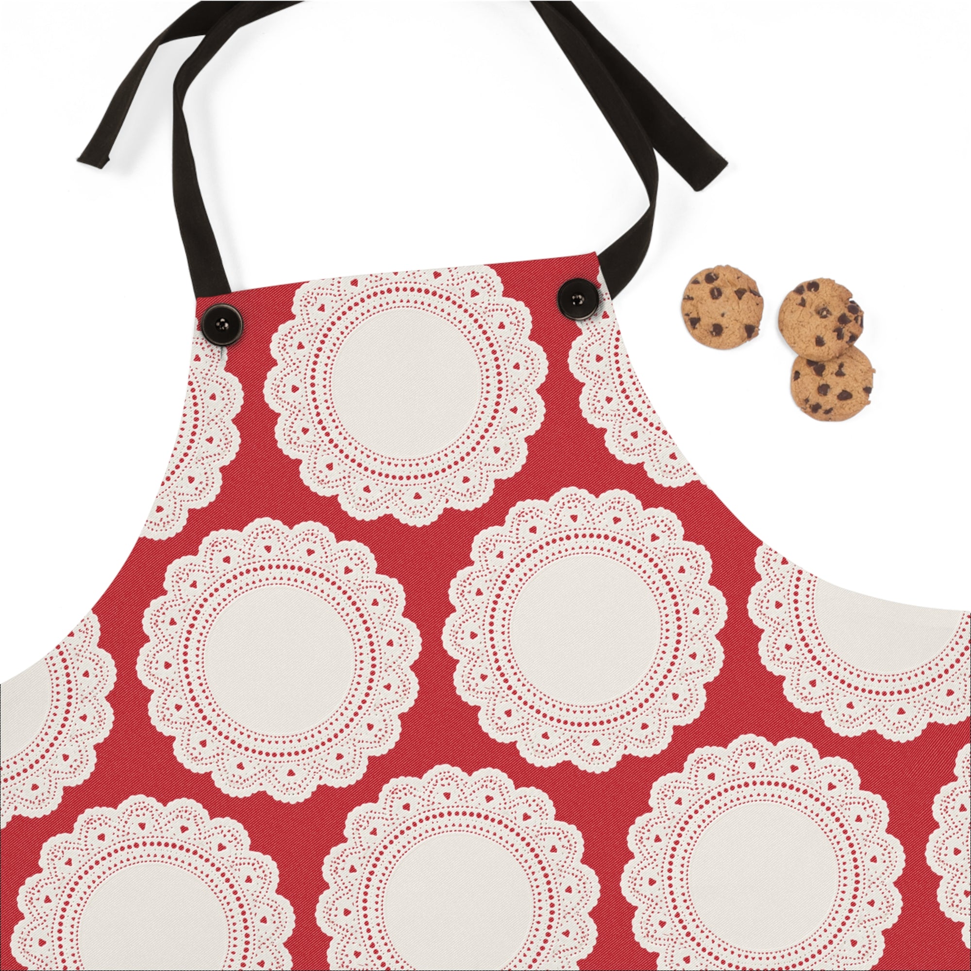The Lace Doily Pattern Poly Twill Apron features a red lace design with a black neck strap, inspired by traditional Polish aprons—an ideal Babcia gift. Three chocolate chip cookies are displayed to the right.