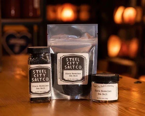Three containers of Black Hawaiian Sea Salt are on a wooden surface with warm lighting: a glass jar, a resealable bag, and a small tin.