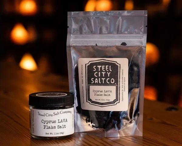 A small jar and resealable bag of Cyprus LAVA Salt, a gourmet finishing salt with activated charcoal, are displayed on a wooden surface with a warmly lit, blurred background.