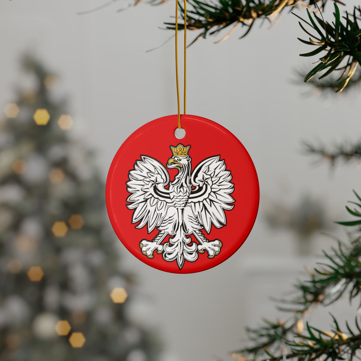 The Polish Eagle Ceramic Ornament, a red circular ceramic piece featuring a white crowned eagle emblem, hangs from a Christmas tree branch, adding elegance and festive charm to your holiday decor.