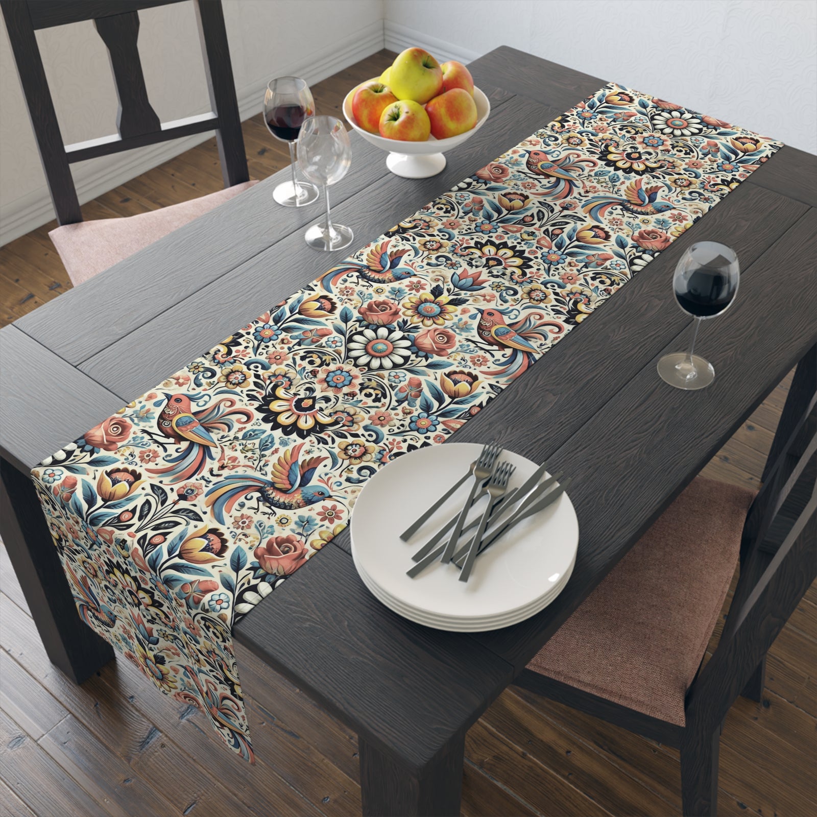 A dark wooden dining table decorated with the Birds and Flowers Floral Table Runner, two wine glasses of red wine, a bowl of assorted fruits, and neatly arranged cutlery on white plates—a charming home decor accent.
