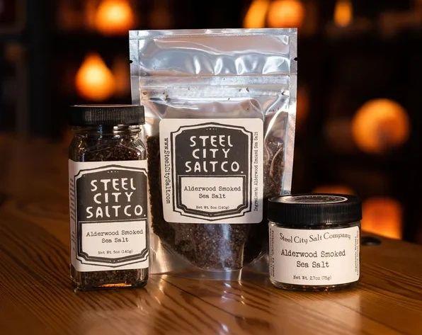 Three containers of Alderwood Smoked Sea Salt—a glass jar, resealable pouch, and small tin—are displayed on a wooden surface with labeled packaging against a warm, blurred background.
