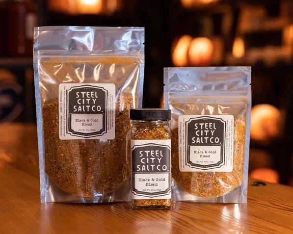Three packages of Black &amp; Gold Blend®—two resealable pouches and one spice jar—are shown on a wooden surface with a softly lit, blurred background.