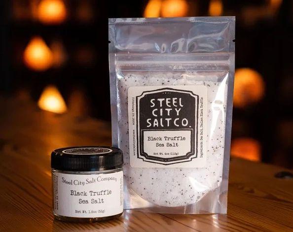 A jar and a resealable bag of Black Truffle Sea Salt rest on wood, featuring rich flavor from Italian black truffles by Steel City Salt Co., while warm, softly blurred lights glow in the background.