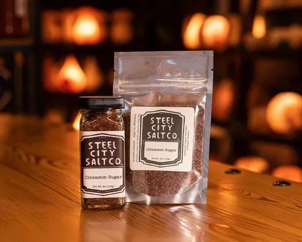 A jar and resealable pouch of Cinnamon Sugar are displayed on a wooden surface with warm lighting, highlighting the rich aroma and flavor of this gourmet blend.