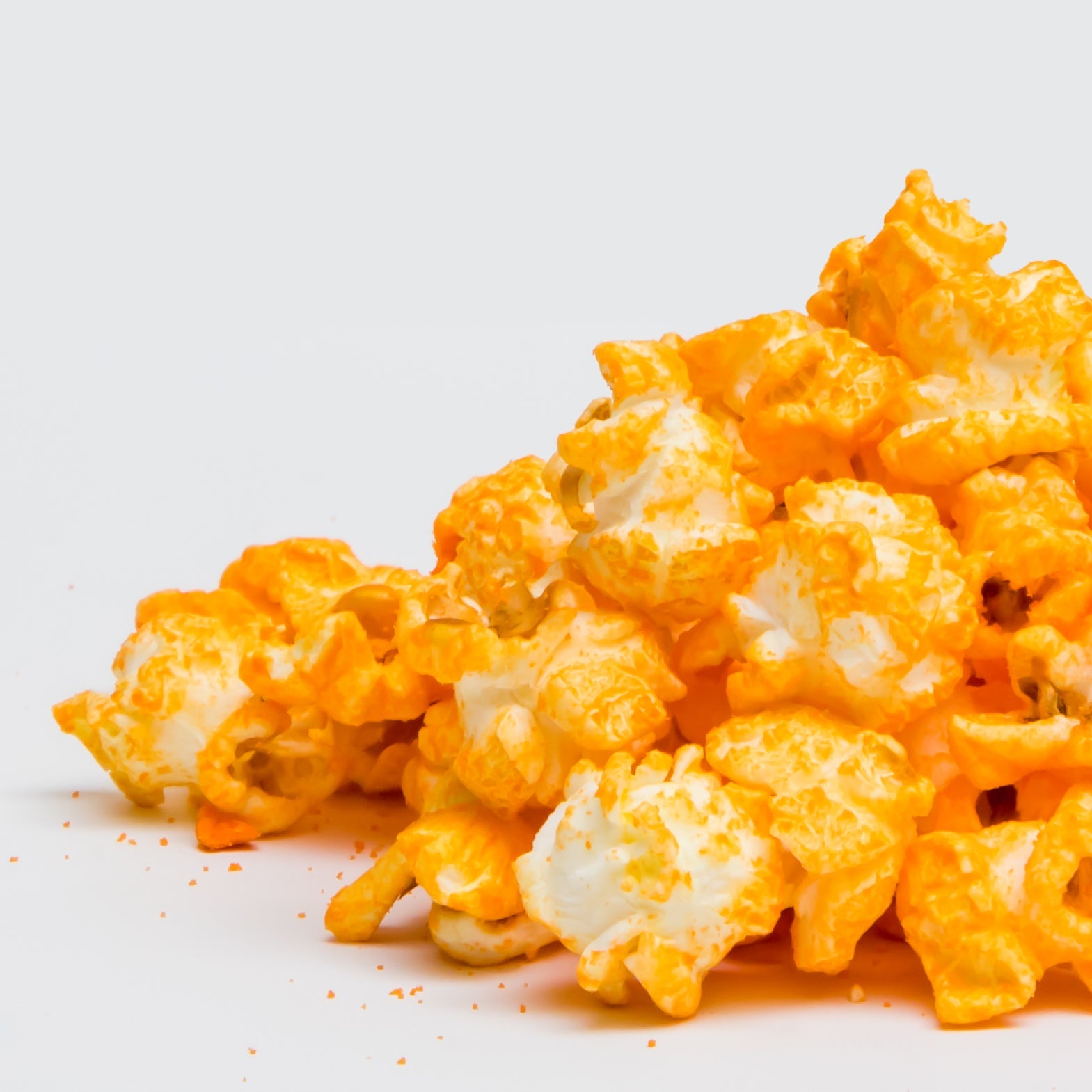 Yellow Cheddar Popcorn is shown in a pile on a white surface, with some real cheddar cheese powder scattered nearby.