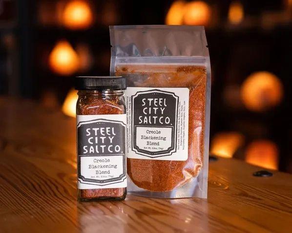 A glass jar and a resealable bag of Creole Blackening Blend sit on a wooden table with a warm, blurred background, ready to add bold Cajun-inspired flavor to your next meal.