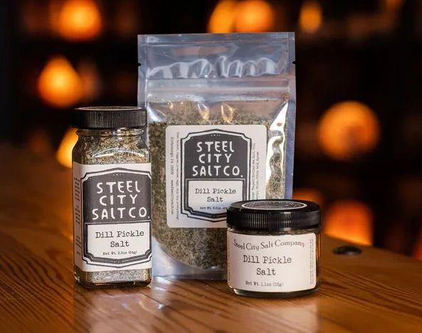 Three containers of Dill Pickle Salt are arranged on a wooden surface with a softly lit background. This tangy blend is available in a jar, pouch, or small tin for adding bold flavor to your favorite dishes.