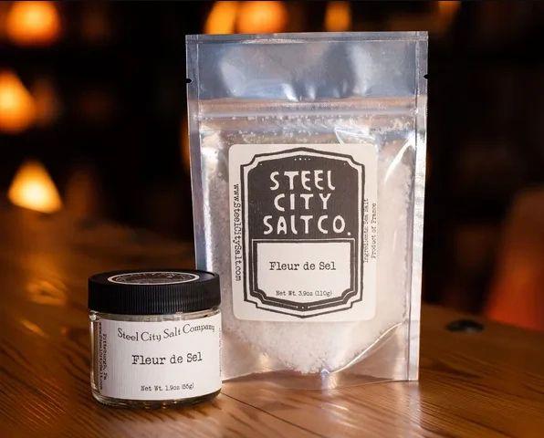 A glass jar and a resealable pouch of Fleur de Sel, a French finishing salt, both featuring black and white labels, sit on a wooden surface with warm, softly blurred lighting in the background.
