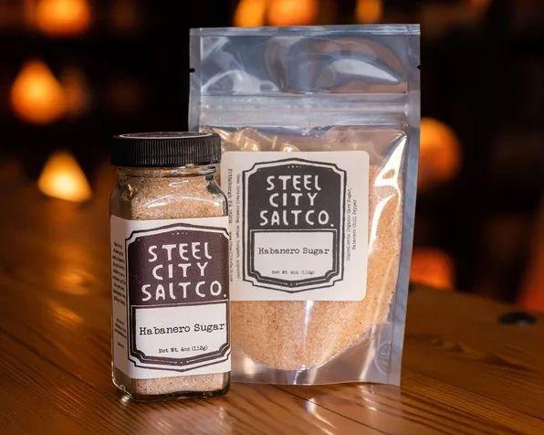 Two containers of Habanero Sugar, a sweet and spicy blend ideal for rimming cocktails, rest on wood: one in a glass jar with black lid, the other in a clear resealable pouch. Warm, blurred lights glow behind them.