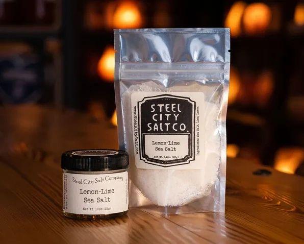 Two containers of Lemon Lime Sea Salt—one a small glass jar, the other a larger resealable bag—sit labeled against a warmly lit background, highlighting the vibrant blend of Pacific sea salt with lemon and lime.