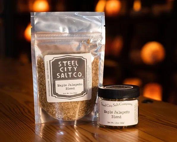 A bag and a small jar of Maple Jalapeno Blend rest on a wooden surface, their gourmet salt glowing in soft, warm light with a gently blurred background.