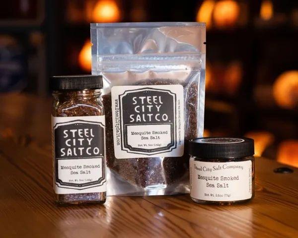 Three containers of Mesquite Smoked Sea Salt—a jar, a resealable pouch, and a small glass jar—sit on a wooden surface, ready to bring bold Texas Barbecue flavor to your next BBQ rub. Warm tones light up the background.