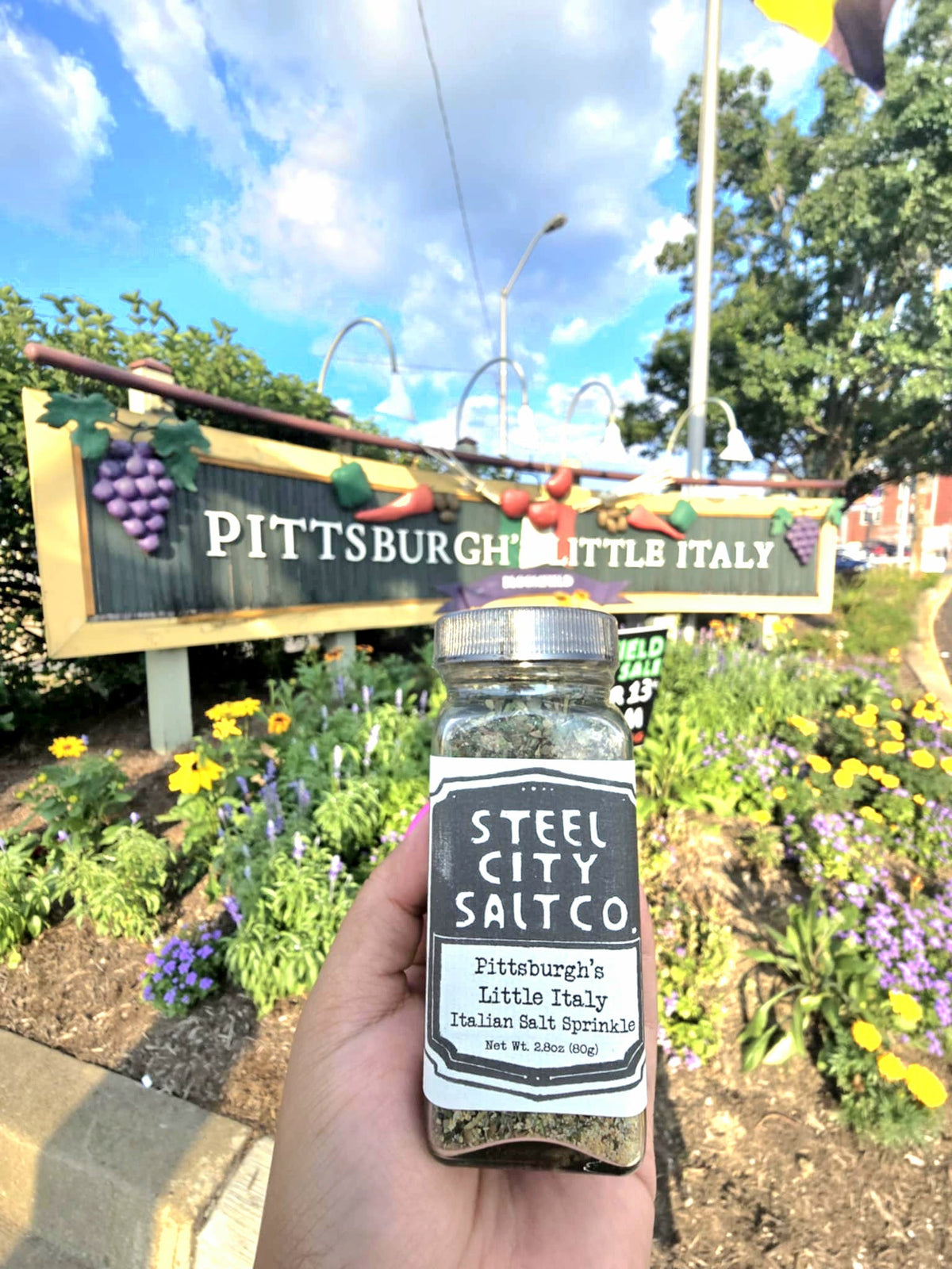 A hand holds a jar of Pittsburgh&#39;s Little Italy Italian Salt Sprinkle before a &quot;Pittsburgh&#39;s Little Italy&quot; sign, with flowers and greenery in the background on a sunny day, highlighting this flavorful all-purpose Italian seasoning.