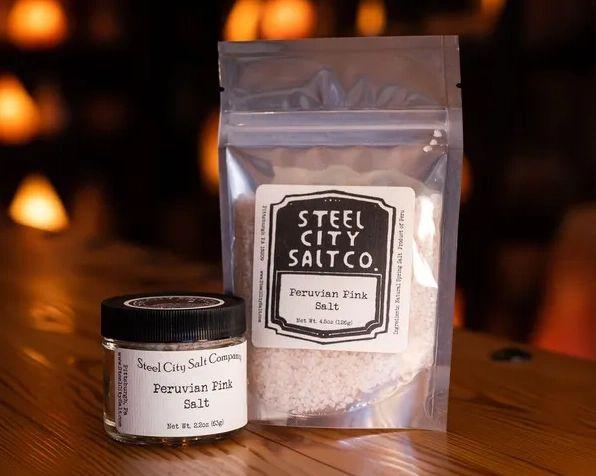 A jar and resealable bag of Peruvian Pink Salt from Steel City Salt Co., ideal as a finishing salt, rest on a wooden surface with a warm, softly blurred orange-lit background.