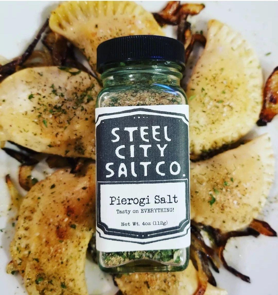 A jar of Pierogi Salt! sits on a plate surrounded by golden-brown Pittsburgh pierogi, topped with herbs and caramelized onions—an ideal way to highlight this savory seasoning blend.