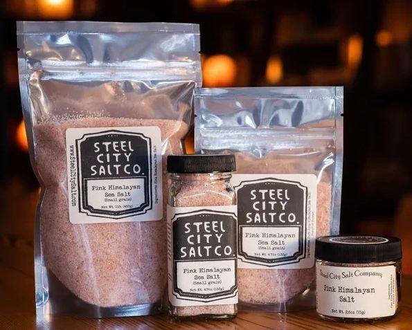 Four containers of Pink Himalayan Salt are shown in different sizes and packaging—resealable bags, a jar, and a tin—arranged on a wooden surface with a warm, blurred background.
