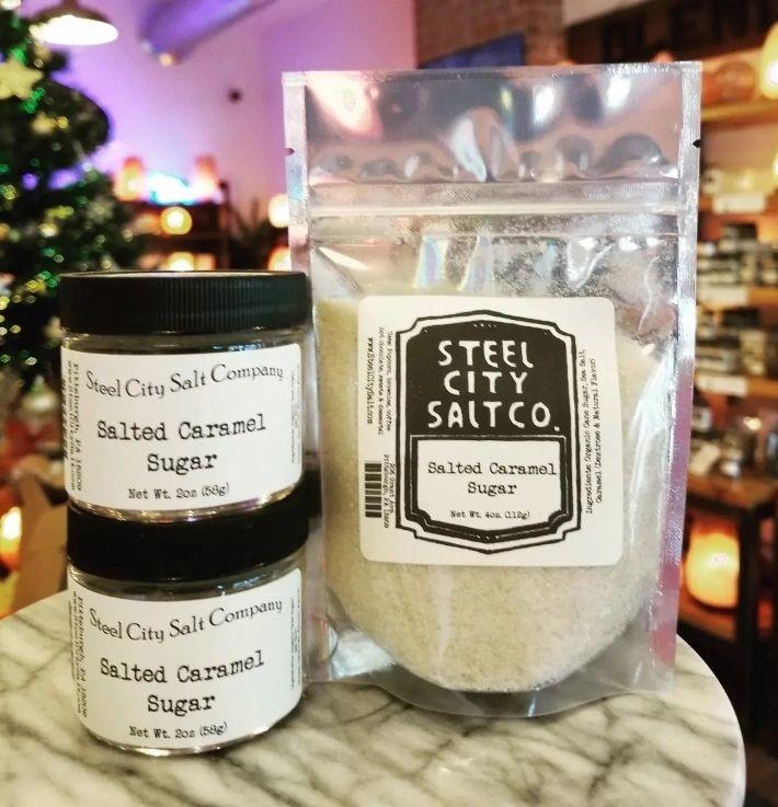 Two small jars and a pouch of Salted Caramel Sugar from Steel City Salt Company, crafted with organic cane sugar, sit on a marble surface. Blurred shelves and a glowing Christmas tree appear in the background.