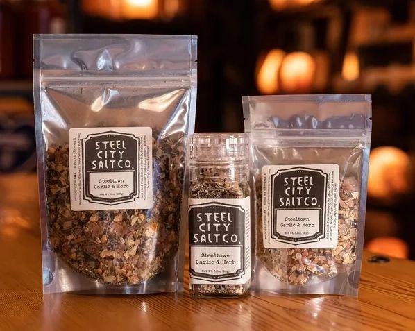 Three packages of Steeltown Garlic & Herb, featuring a Himalayan salt grinder and two resealable bags, are arranged on a wooden surface under warm lighting for a cozy feel.