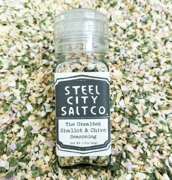 A bottle of &quot;The UnSalted Shallot &amp; Chive Seasoning (Salt Free)&quot; rests on a surface sprinkled with colorful dried flakes, highlighting this vibrant, all-purpose herb blend as a flavorful salt-free seasoning choice.