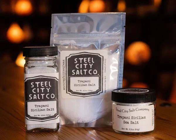 Three containers of Trapani Sicilian Sea Salt—glass jar, resealable bag, and plastic jar—are shown on a wooden surface with a warm, blurred background.