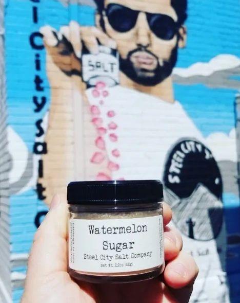 A hand holds a jar of Watermelon Sugar—ideal for dessert rims—in front of a mural of a man sprinkling salt, set against bold blue and white tones.