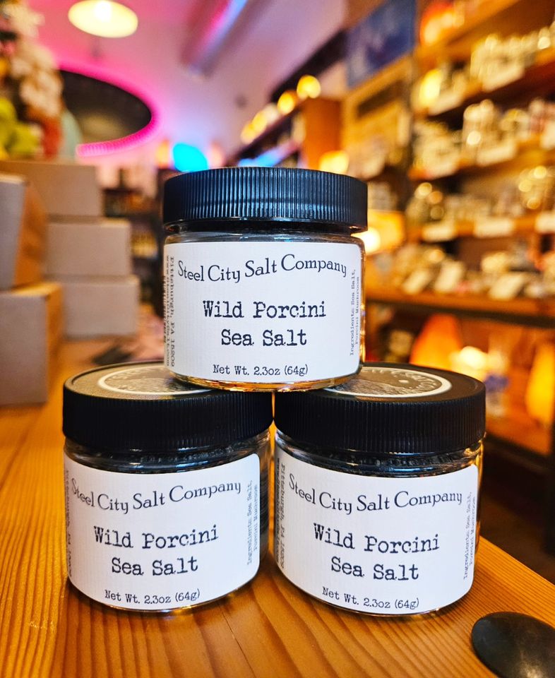 Three jars of Wild Porcini Salt are stacked on a wooden counter in a warmly lit, cozy shop filled with shelves stocked with various products.