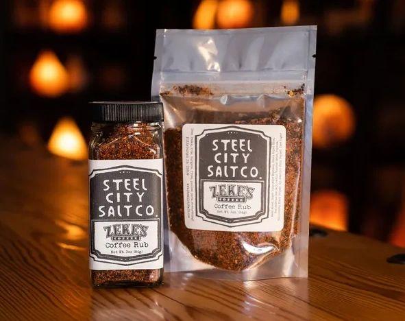 A glass jar and resealable pouch of ZEKE's Coffee Rub with Red Hawaiian Sea Salt rest on a wooden surface against a warmly-lit, blurred background.