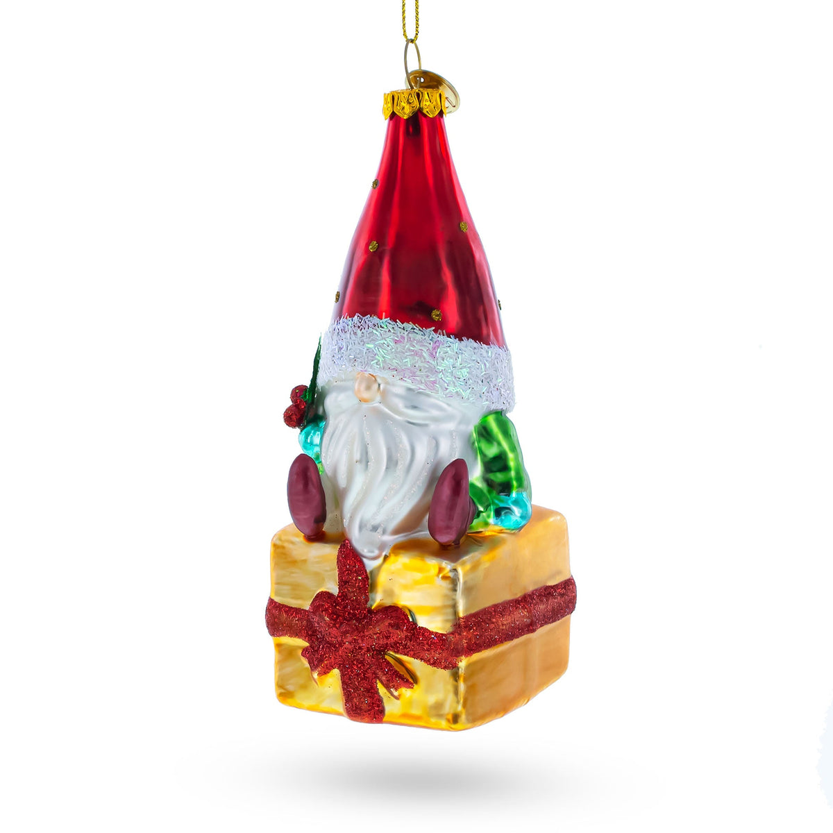 The Gnome Bearing Gifts Glass Christmas Ornament features a festive gnome in a tall red hat and green outfit, sitting on a gold present with a glittery red bow, hanging by a gold string against a white background.